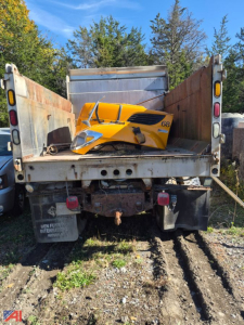 Dump truck bed rear view with a yellow truck cab shell in the flatbed and dirt and grass on ground.