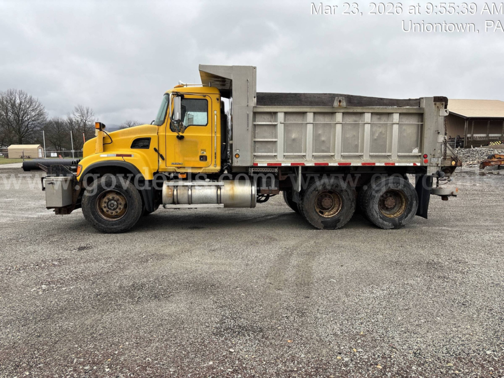 Dump truck side view showing a yellow Mack CV713 with a large metal dump bed and dual rear axles.