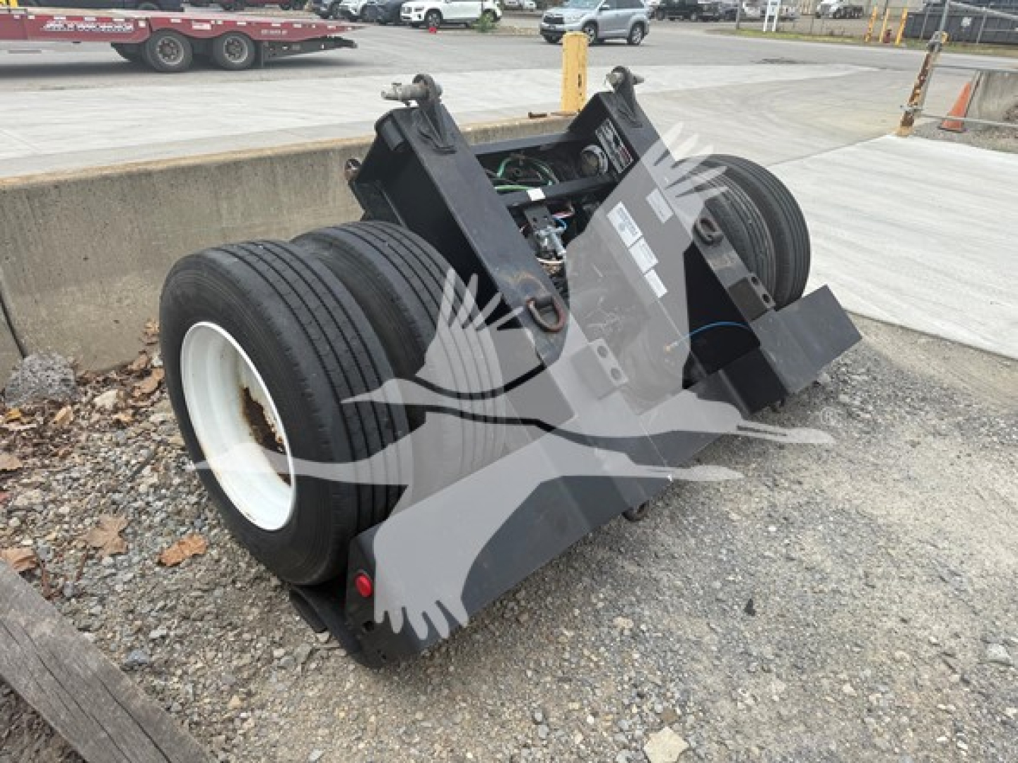 Talbert flip axle trailer component showing dual wheels and mounting assembly with hydraulic lines and labels, lot and equipment in background.