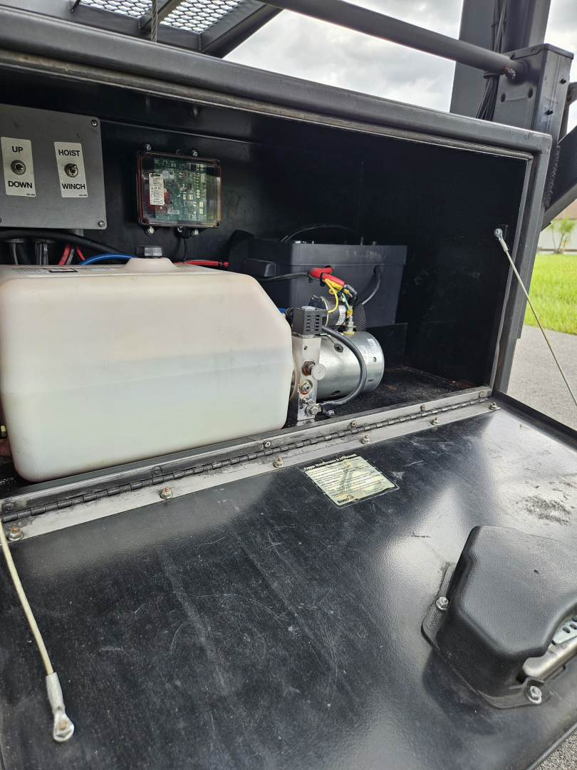 Truck equipment bay showing a white polyethylene reservoir and small electric motor with wiring in a black enclosure under a Dodge Ram body. Labels and control electronics visible.