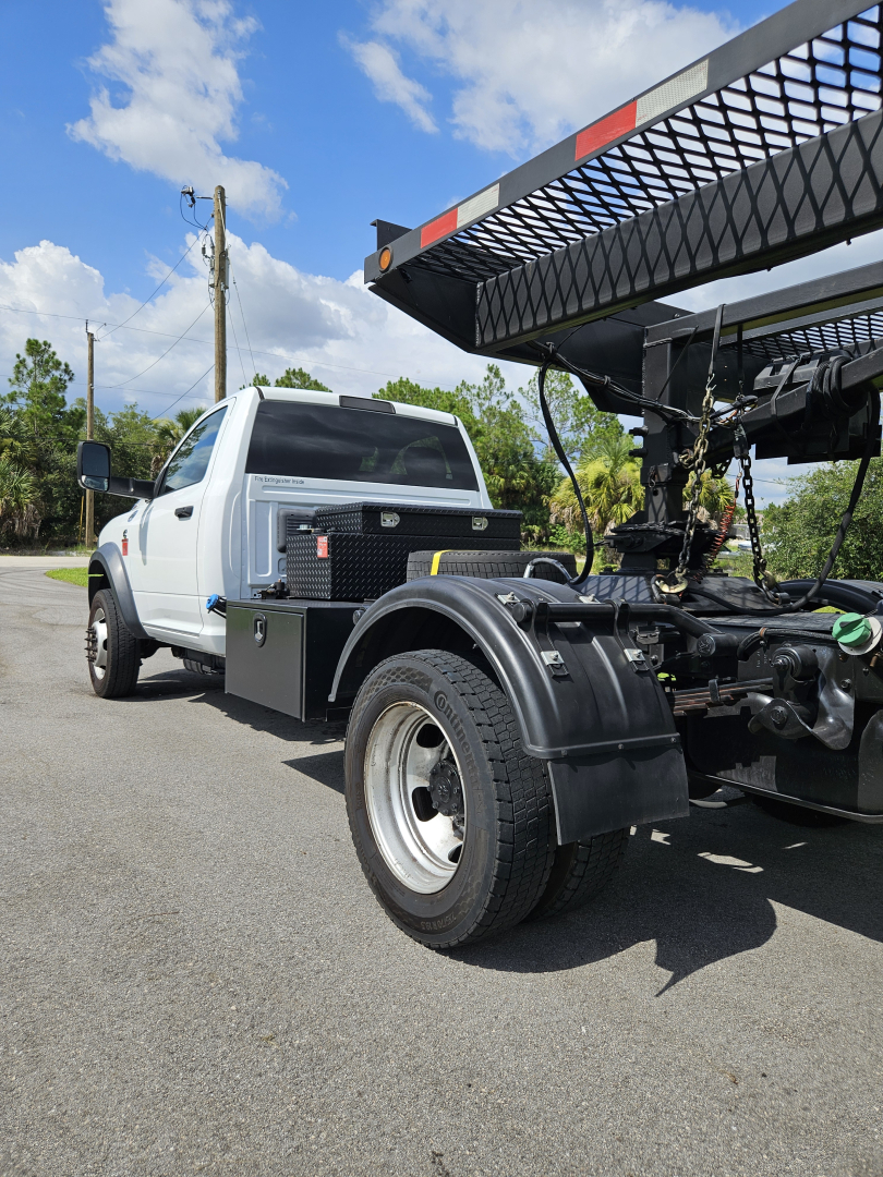 Partial side view of a white Dodge Ram truck with a large black hydraulic flatbed/metal rack attachment and utility toolbox visible on the side.