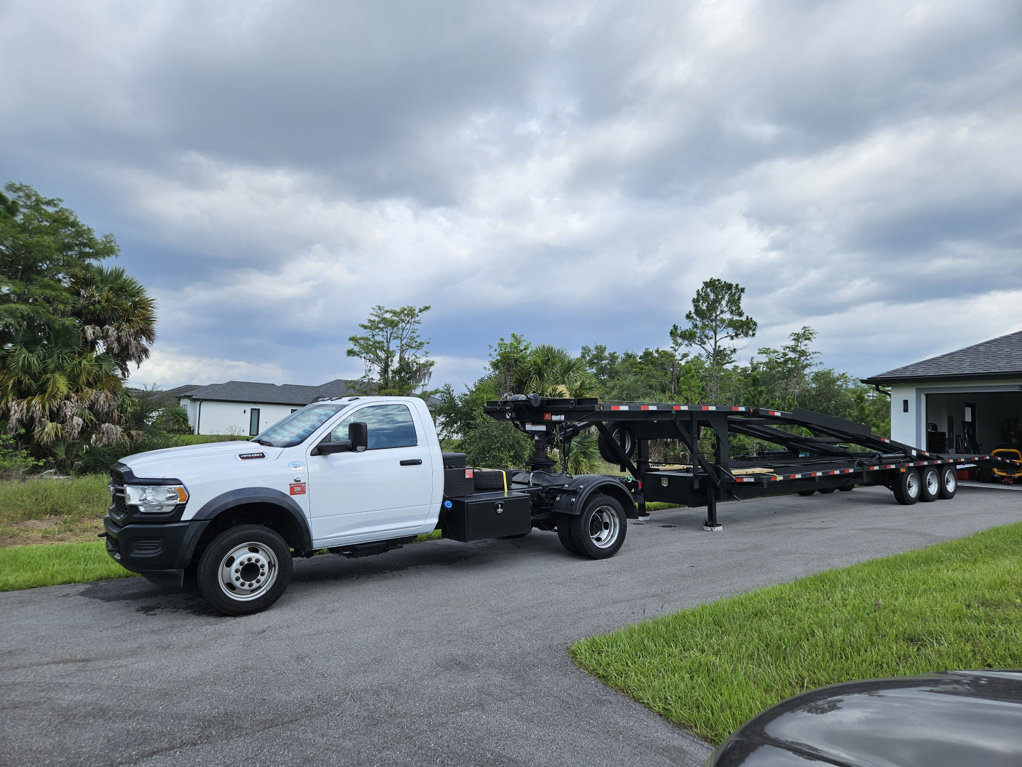 Truck with a white Ram 5500 chassis cab and a long car trailer, displayed in a driveway with a cloudy sky.