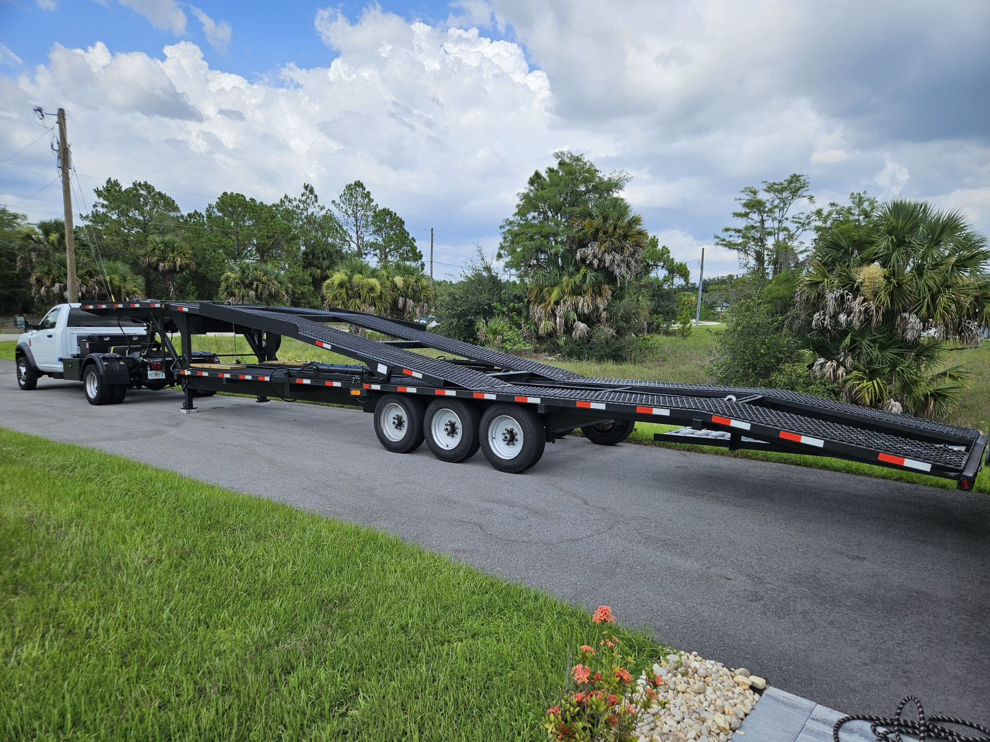 Flatbed tow trailer attached to a white Dodge Ram pickup truck, long multi-axle equipment trailer on a driveway with trees and blue sky