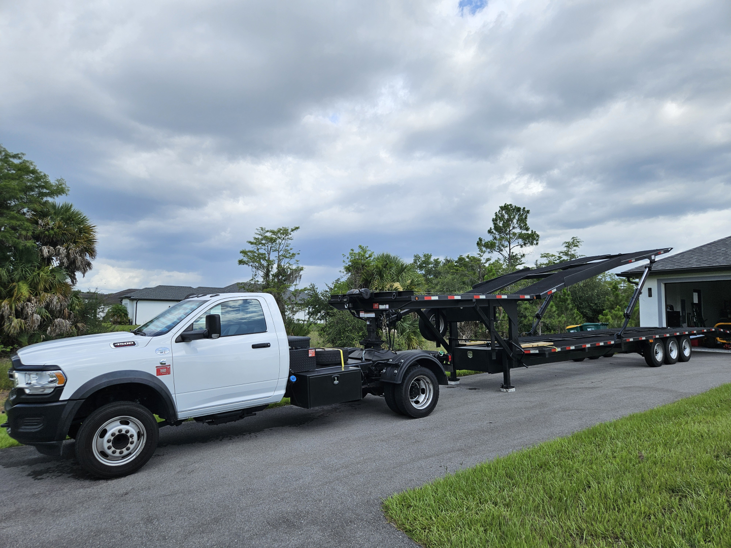 White Ram pickup truck with a long flatbed trailer on a tow truck, viewing from the passenger side, with a cloudy sky and grassy area in the background.