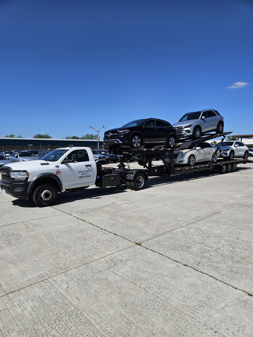 Truck carrier with a white tow truck trailer transporting multiple Dodge Ram vehicles on a car hauler under a blue sky.