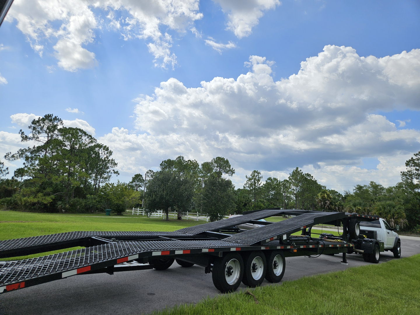 Truck with a car hauler trailer attached, extended ramp sections in a grassy area under a blue sky with clouds.