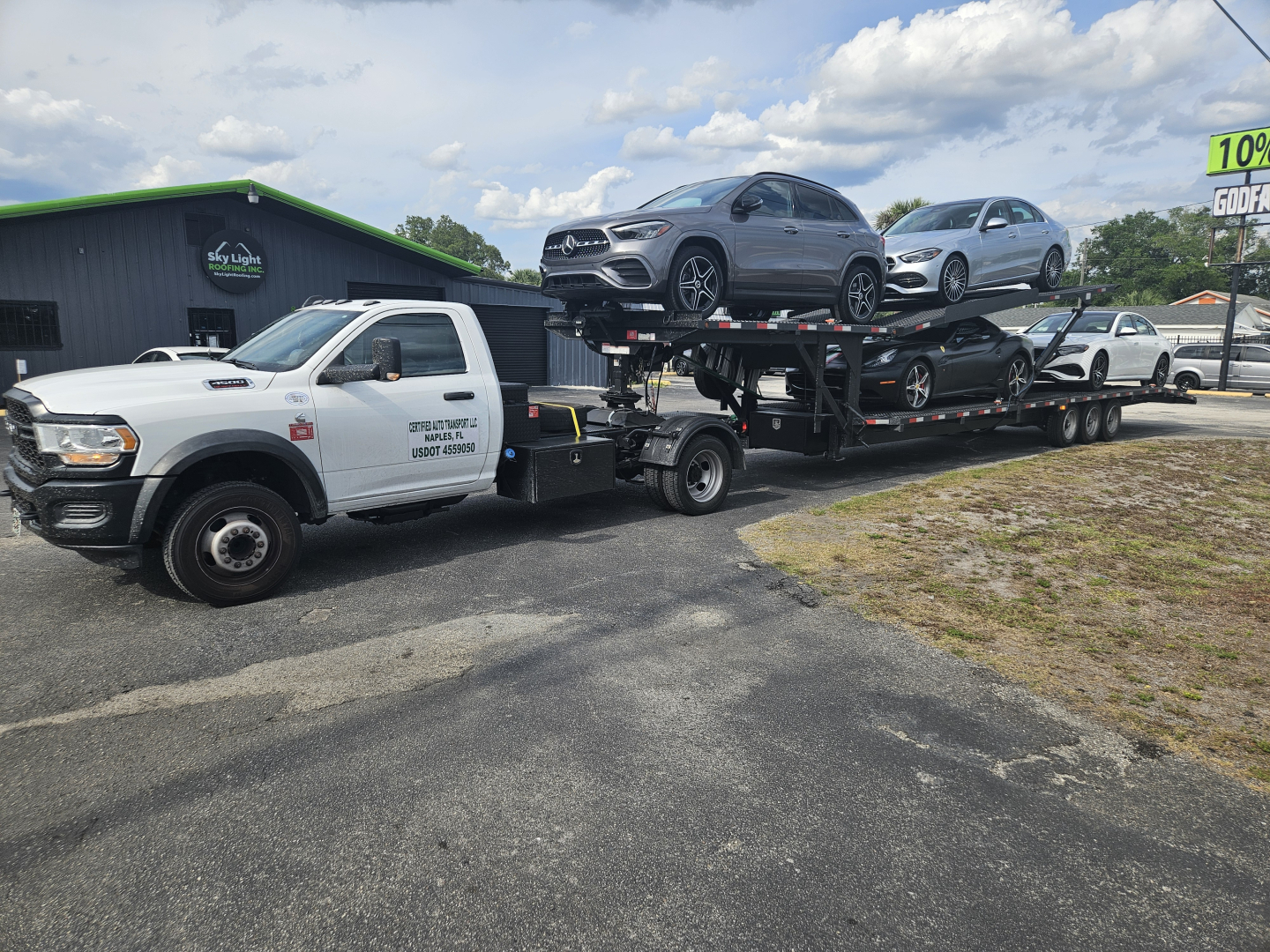 Truck with car carrier trailer transporting two SUVs, white pickup truck towing on trailer, dealership lot in background with sign and green building.