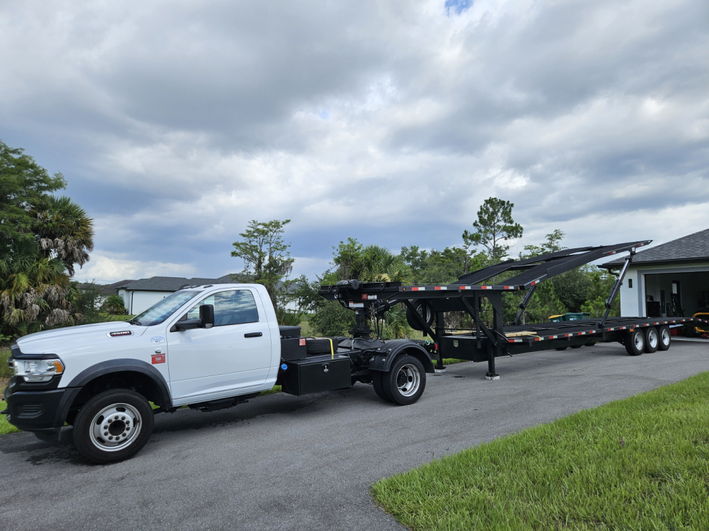 White Ram pickup truck with a long flatbed trailer on a tow truck, viewing from the passenger side, with a cloudy sky and grassy area in the background.