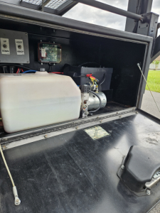 Truck equipment bay showing a white polyethylene reservoir and small electric motor with wiring in a black enclosure under a Dodge Ram body. Labels and control electronics visible.