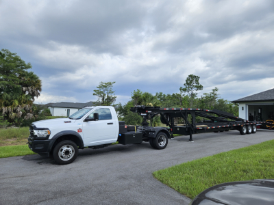 Truck with a white Ram 5500 chassis cab and a long car trailer, displayed in a driveway with a cloudy sky.