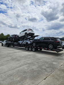 Car carrier trailer with two levels transporting multiple vehicles; white SUV on top, other cars visible on the lower level.