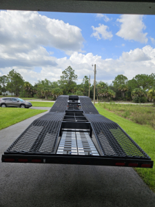 Flatbed trailer with metal grated surface and ramps, viewed from the rear, open lane down center with winch and mounting points.
