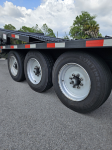 Truck trailer wheels and rims with multiple axles, tire sidewalls visible; part of a flatbed trailer frame shown.