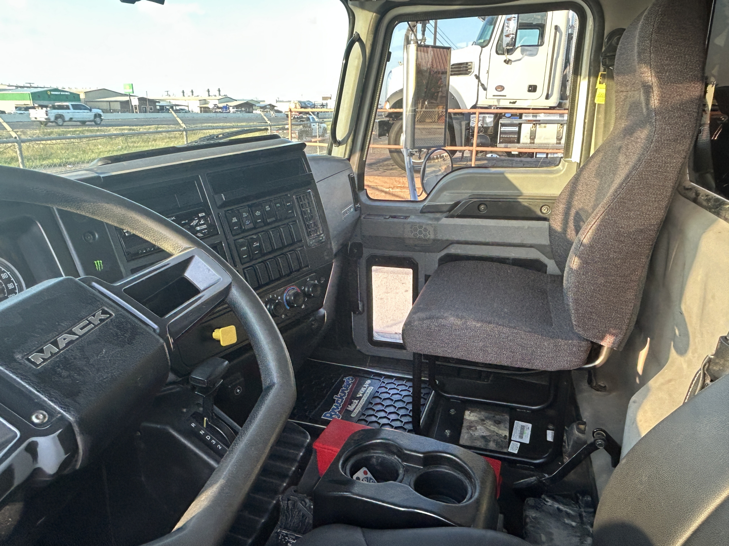 Truck cab interior showing steering wheel with 'MACK' logo, dashboard controls, driver seat, center console with cup holders, and front passenger area.