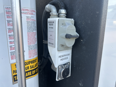 Reservoir heater switch panel and hoses in engine bay of a 2021 Mack MD642 service truck, with warning labels and fluid lines visible.