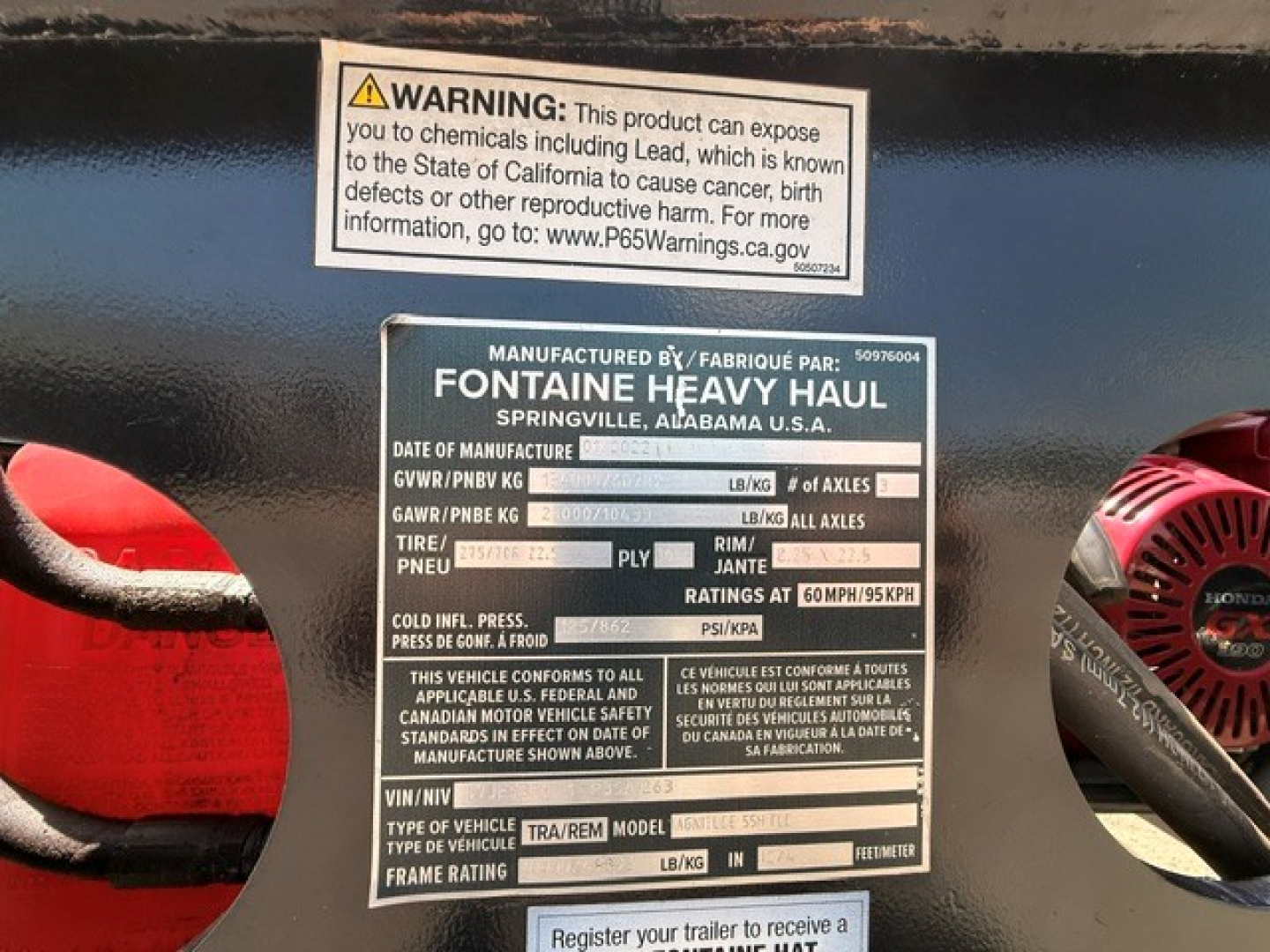 Label plate on Fontaine Heavy Haul trailer showing manufactured by Fontaine; text lines with GVWR/PNBV, tire, rim, and axle ratings; warning sticker above.