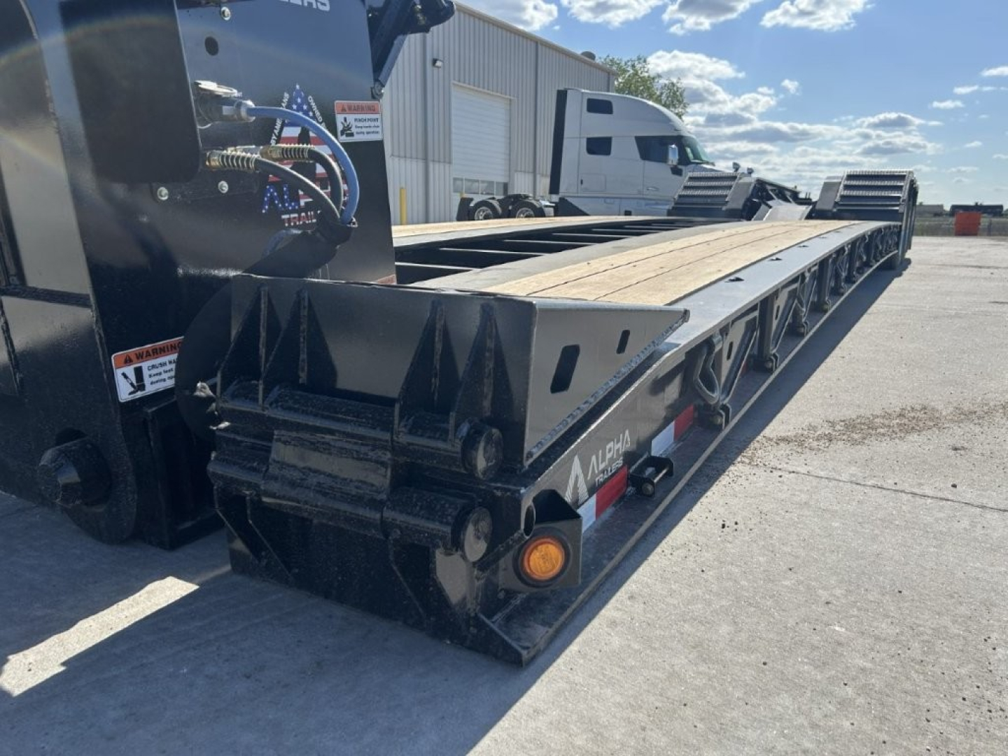 Lowboy trailer showing hydraulic components and long wooden deck with multiple axles under the frame. Visible labels include Alpha A110HDG-SF-18" LDH branding on the side and metal ramp sections at the rear.