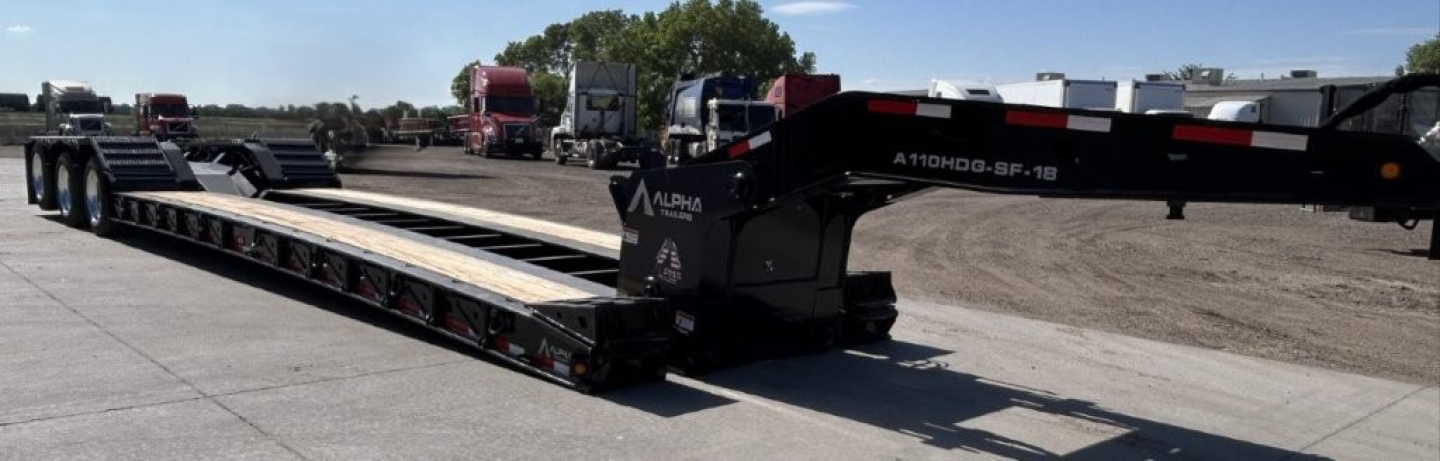 Long lowboy trailer in a yard showing A110HDG-SF-18 text on the gooseneck with deck extending, multiple axles, and graphics.