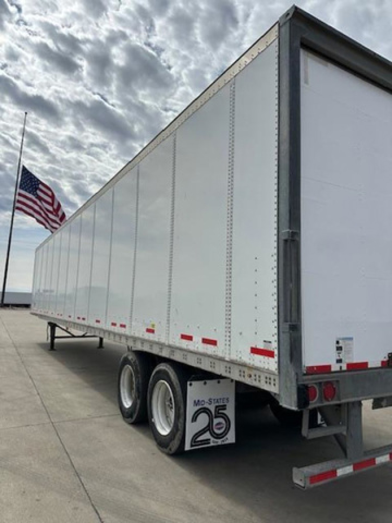 Trailer side view showing white Wabash van trailer with dual rear axles and Mo-State 25 logo mudflap; US flag in background.