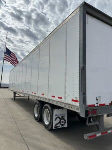 Trailer side view showing white Wabash van trailer with dual rear axles and Mo-State 25 logo mudflap; US flag in background.