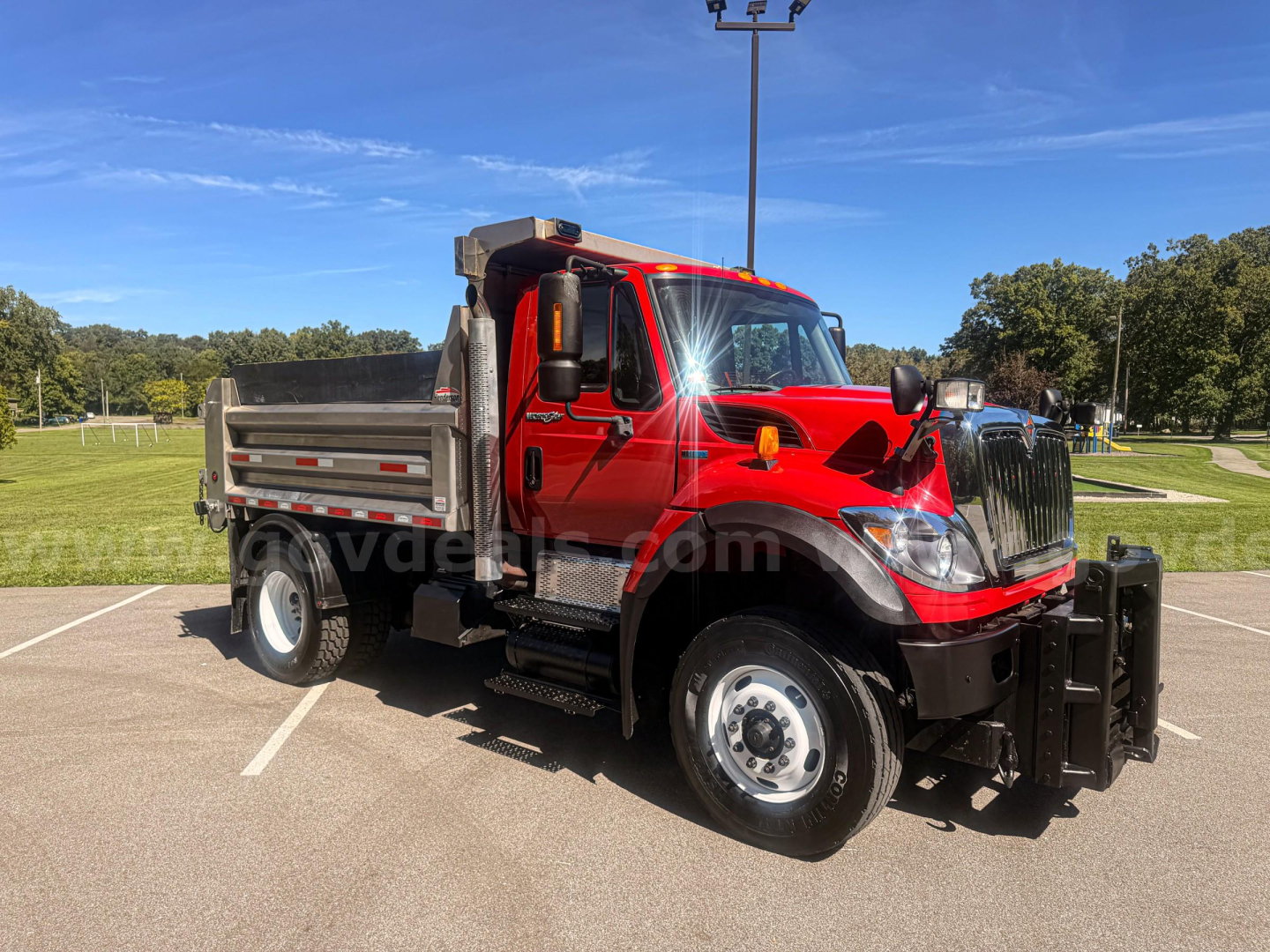 Truck cab in red with dump bed and plow equipment, International 7400 series, side view showing front bumper and wheel, plow mount and guard visible.