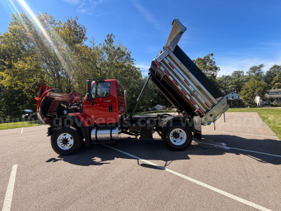 Truck bed tilted dumping with red International 7400 cab and hydraulic system visible, plow-ready dump truck in parking lot.