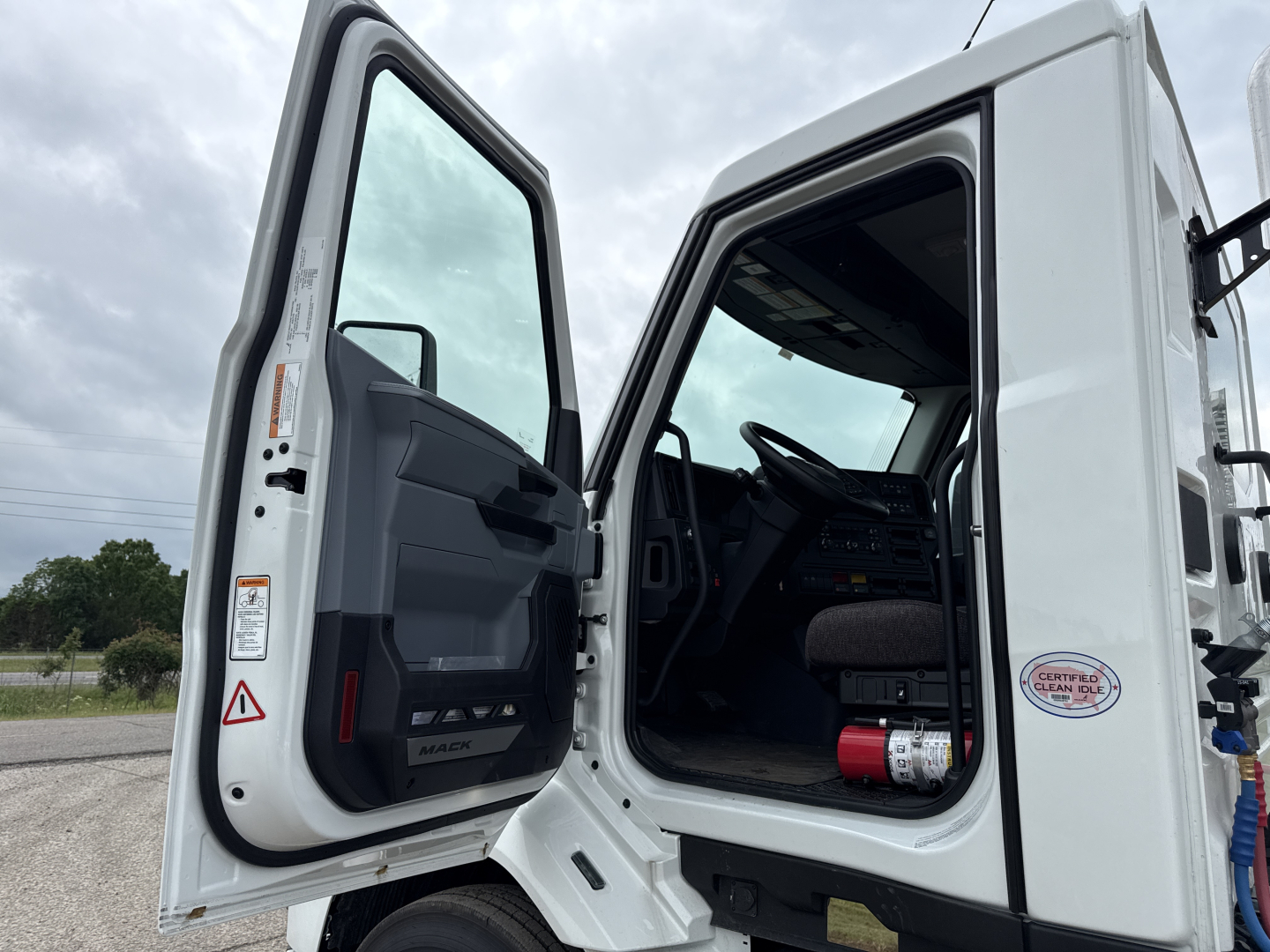 Truck cab interior with open doors showing steering wheel, dashboard, and steps; white 2027 Mack AN64T conventional daycab data plate visible on door frame