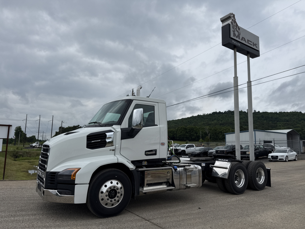 Mack AN64T truck tractor in a dealership lot, white cab, side tanks, three rear axles with dual wheels, Mack sign in background.