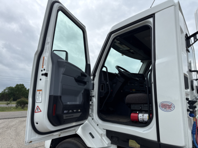 Truck cab interior with open doors showing steering wheel, dashboard, and steps; white 2027 Mack AN64T conventional daycab data plate visible on door frame