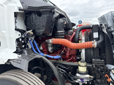 Truck engine bay of a new 2027 Mack AN64T with exposed red engine, orange coolant hose, blue lines, radiator, and various components visible.