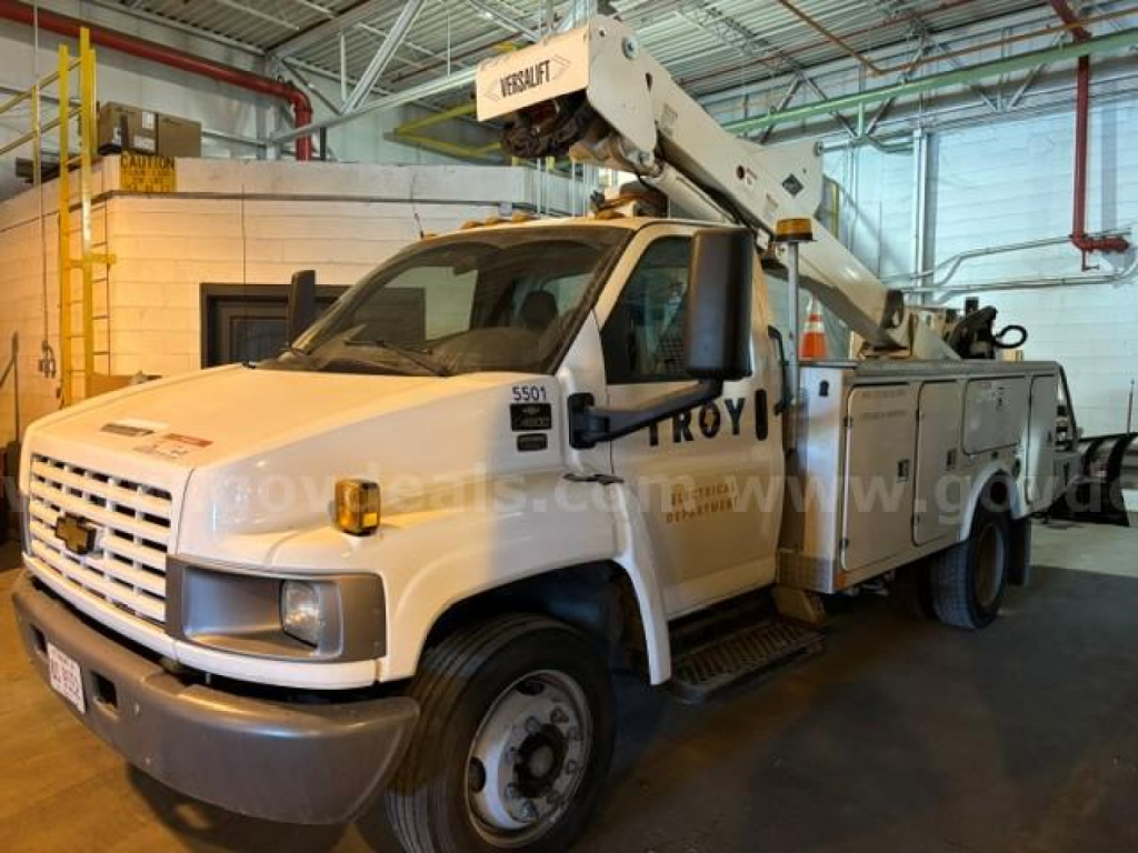 Truck in service body with bucket lift labeled 'US LIFT' and utility compartments, white Chevrolet truck with boom on top, indoor showroom.