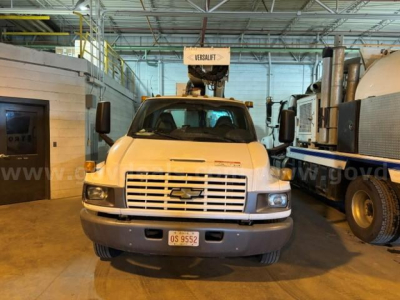 Front view of a white Chevrolet truck in a warehouse setting with a mounted Versalift bucket on the cab roof and a large industrial vehicle to the right.