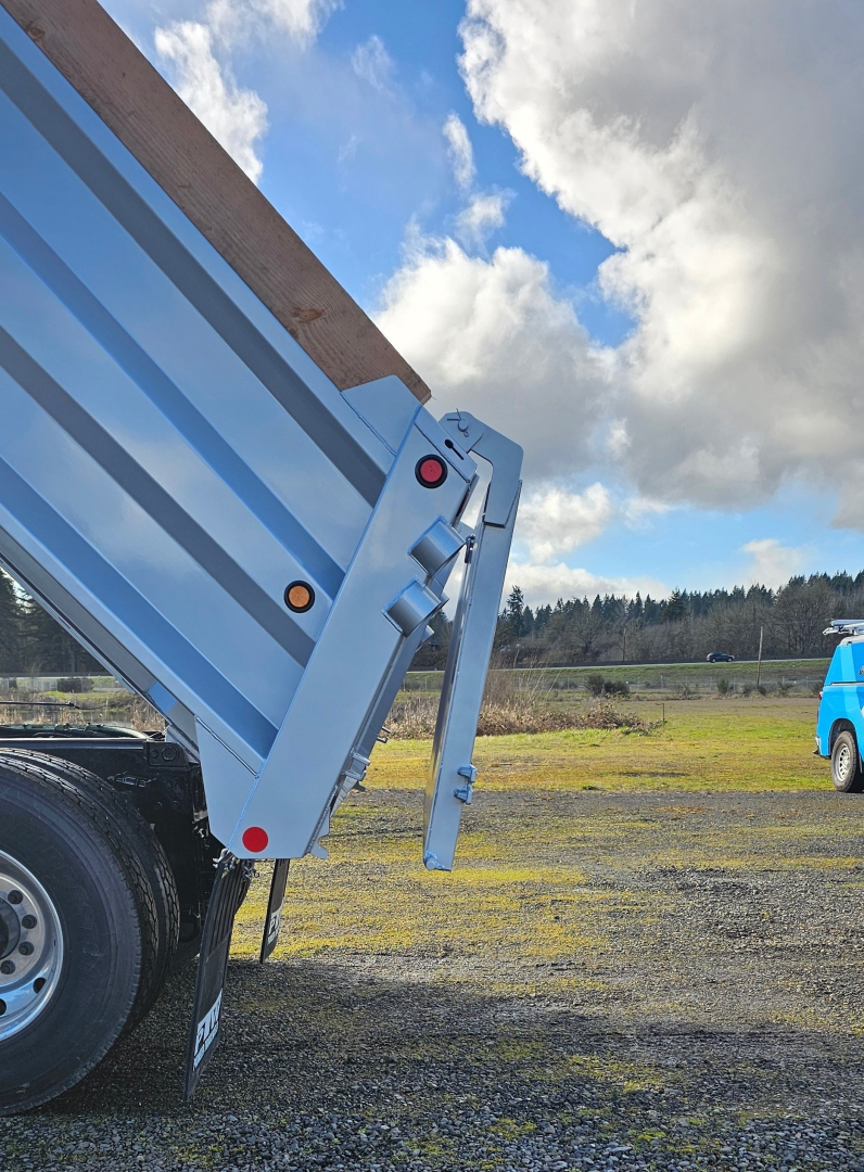 Dump truck bed side showing hydraulic tailgate hinge and blue trailer, with gravel ground and sky in background.