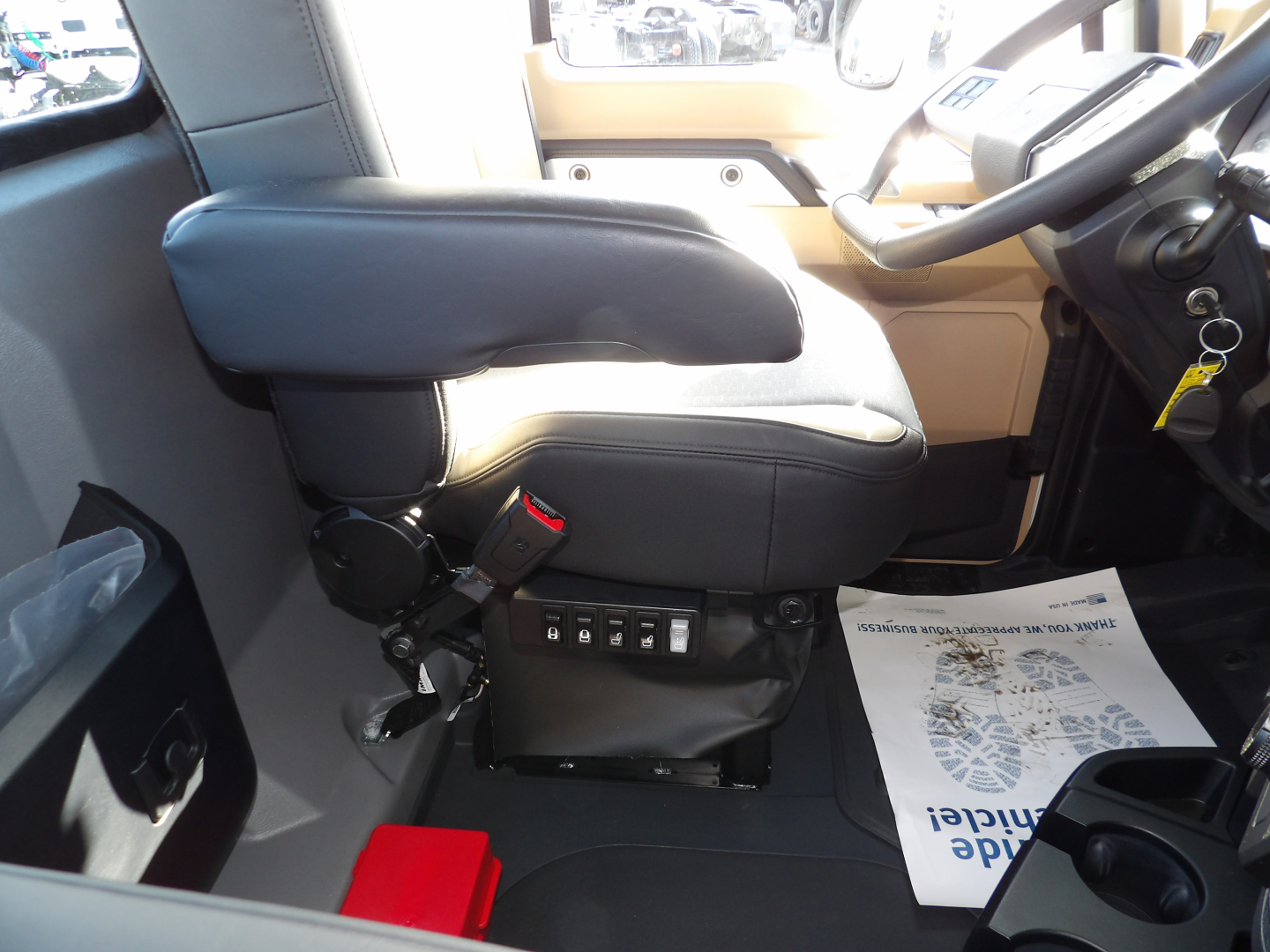Cab interior of a new Mack GR104F dump truck showing driver’s seat with adjustable armrest and control switches beneath, steering wheel, and floor mat visible.