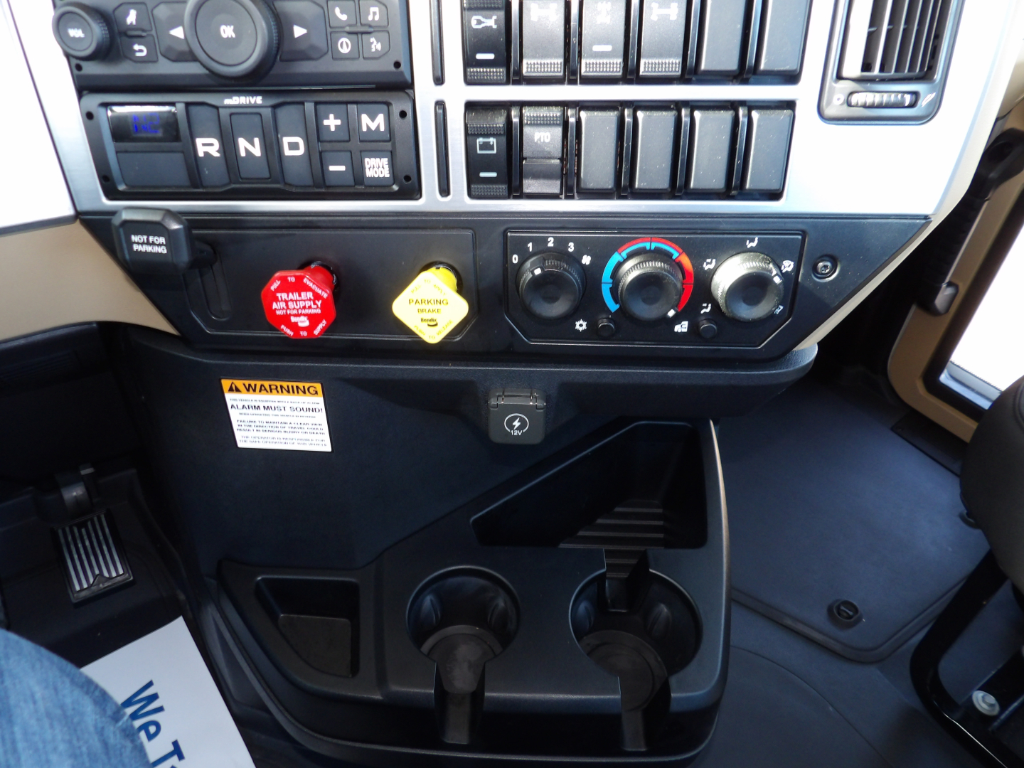 Dashboard of a Mack GR104F truck showing gear selector R N D plus auxiliary buttons, climate controls, two cupholders, warning label, and pedal area.