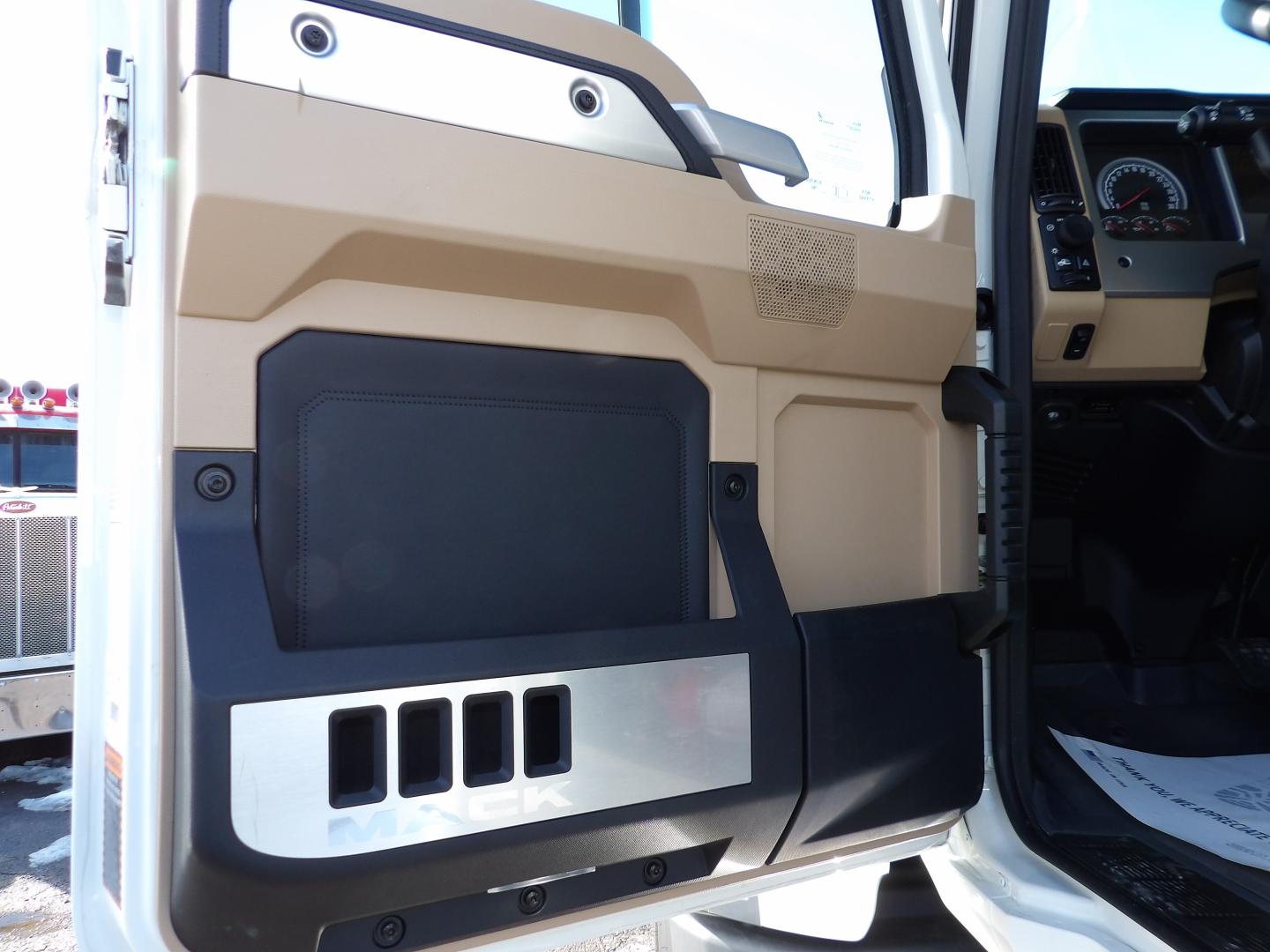 Truck door panel interior of a 2025 Mack GR104F dump truck showing beige plastic and black storage pocket with metal accent; part of dashboard and steering wheel visible.