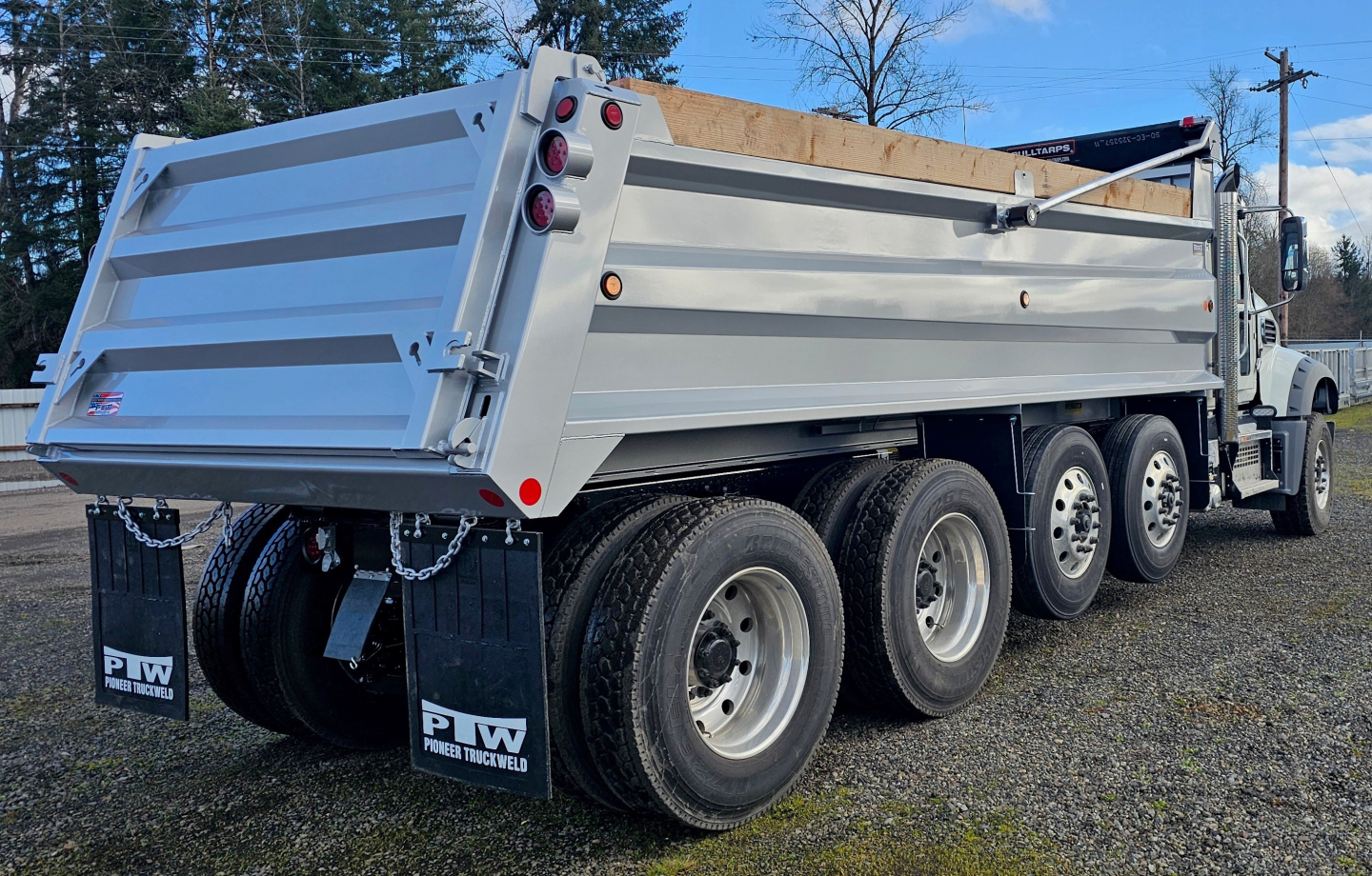 Dump truck bed with three rear axles showing light-gray aluminum dump body and dual rear tires.