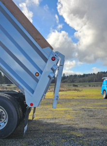 Dump truck bed side showing hydraulic tailgate hinge and blue trailer, with gravel ground and sky in background.