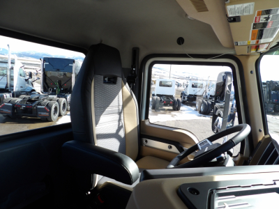 Cab interior of a new 2025 Mack GR104F dump truck showing steering wheel, driver's seat, and dashboard with warning stickers; exterior lot with other trucks visible through window.