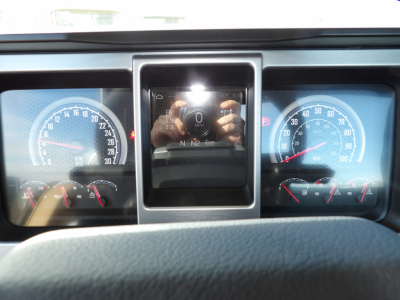 Vehicle dash panel showing two round gauges (left tachometer and right speedometer) with smaller gauges below; center digital display; reflection of photographer in screen.