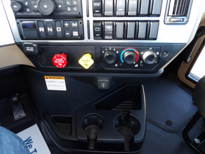 Dashboard of a Mack GR104F truck showing gear selector R N D plus auxiliary buttons, climate controls, two cupholders, warning label, and pedal area.