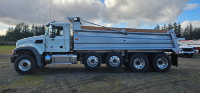 Dump truck side view showing Mack GR104F with a large dump body and five rear axles, chrome fuel tank, and California-style cab; no visible text discernible on the truck from this angle.