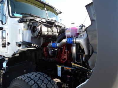 Front left engine bay of a 2025 Mack GR104F dump truck showing hoses, intake, and coolant reservoir; visible components include turbo/intercooler piping and engine block.