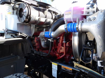 Truck engine bay showing Mack GR104F diesel engine with blue hoses, coolant reservoir, and pink DEF/overflow bottle visible.