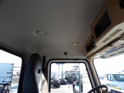Interior of a new Mack GR104F dump truck cab showing overhead storage compartments and windshield view, with other trucks visible outside.