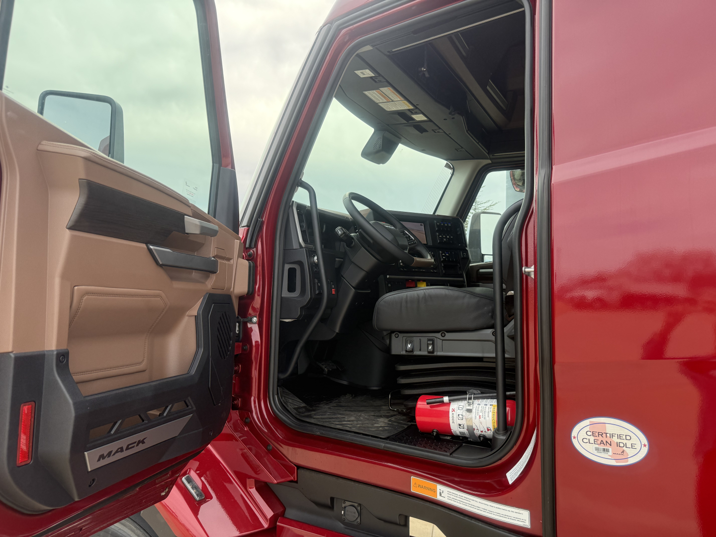 Truck cab interior view through open driver door showing steering wheel and dashboard of a red 2026 Mack PR64T76H, with a red fire extinguisher mounted near the door sill.