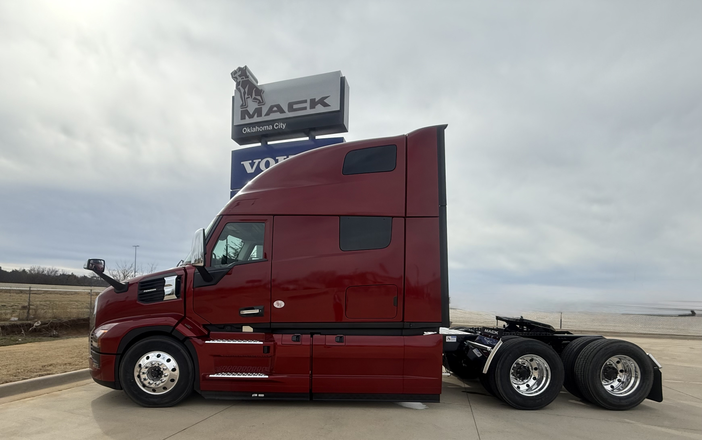 Truck side view of a red 2026 Mack PR64T76H conventional sleeper, long cab with aerodynamic mirrors, parked on pavement near a Mack sign.