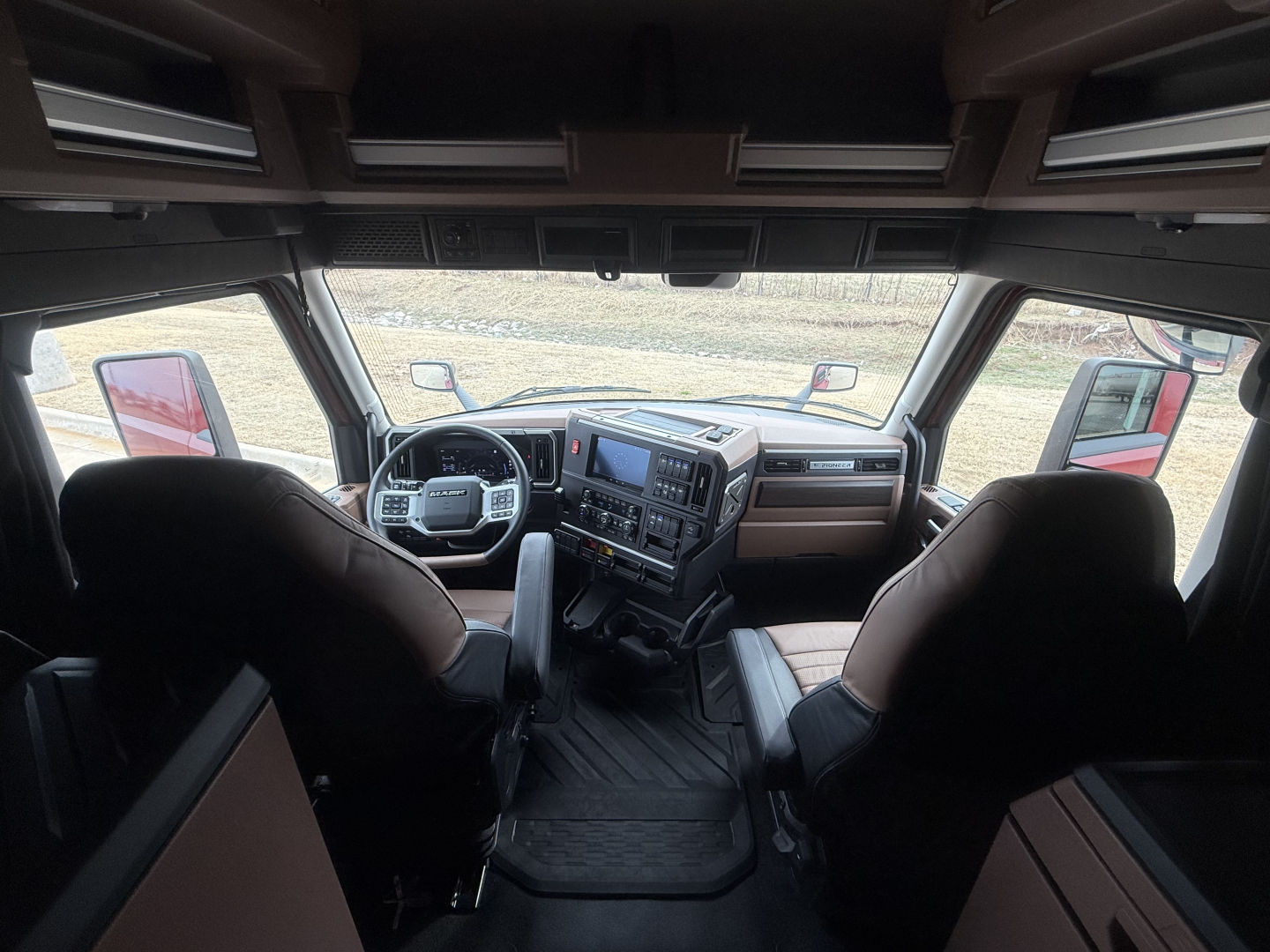 Interior of a new Mack PR64T76H conventional sleeper truck cab, driver and passenger seats, dashboard with large infotainment screen, center console, steering wheel, and overhead storage visible.
