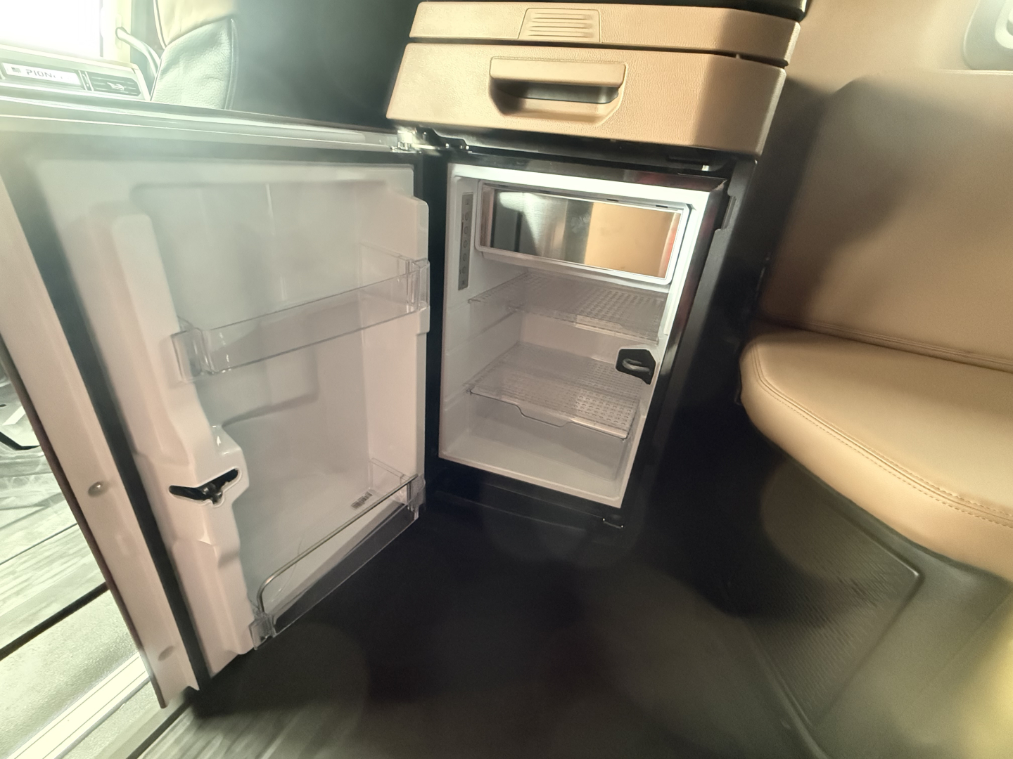 Interior of a 2026 Mack PR64T76H showing a compact under-dash refrigerator with open doors revealing shelves and a small freezer compartment.