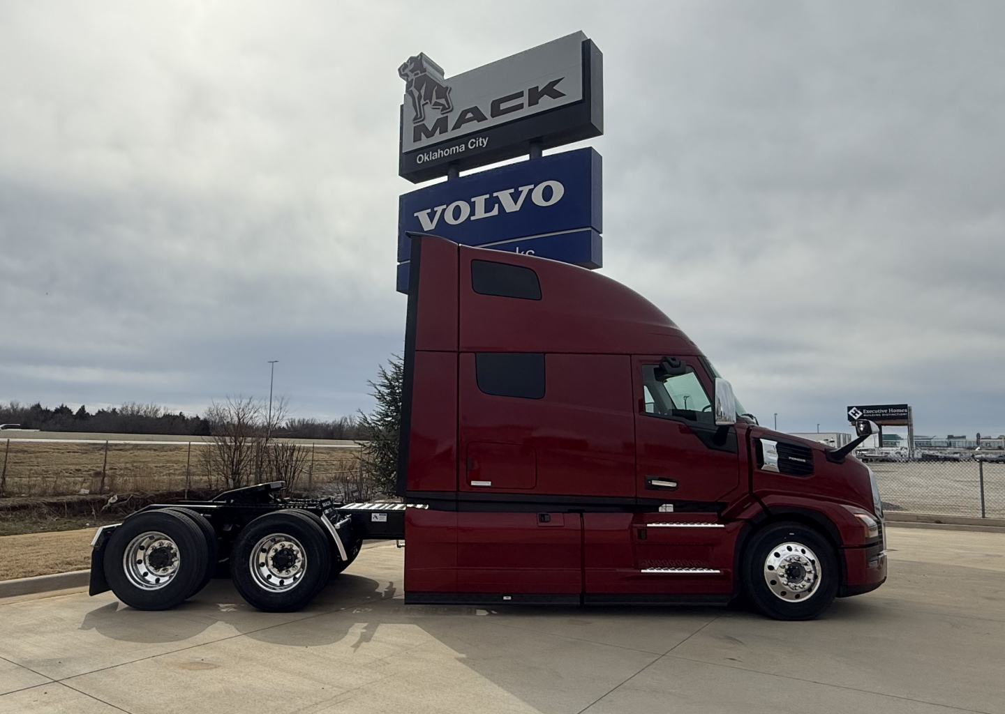 Truck side view showing red 2026 Mack PR64T76H conventional sleeper with 6x4 chassis and Volvo dealership sign in background.